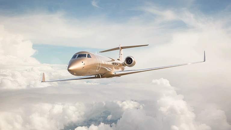 A Gulfstream G550 from JetWise soars above a sea of clouds with a clear blue sky as its backdrop. The jet’s sleek, metallic fuselage reflects light, highlighting its streamlined design as it navigates through the high-altitude atmosphere.